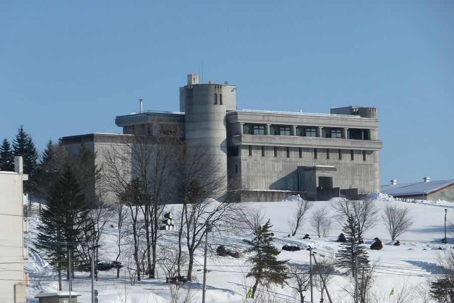 雪に覆われた丘の上に建つ、円筒形の塔をもつ灰色のコンクリートの大きな建物。手前に葉を落とした木々と針葉樹、雪の斜面に積まれた樽