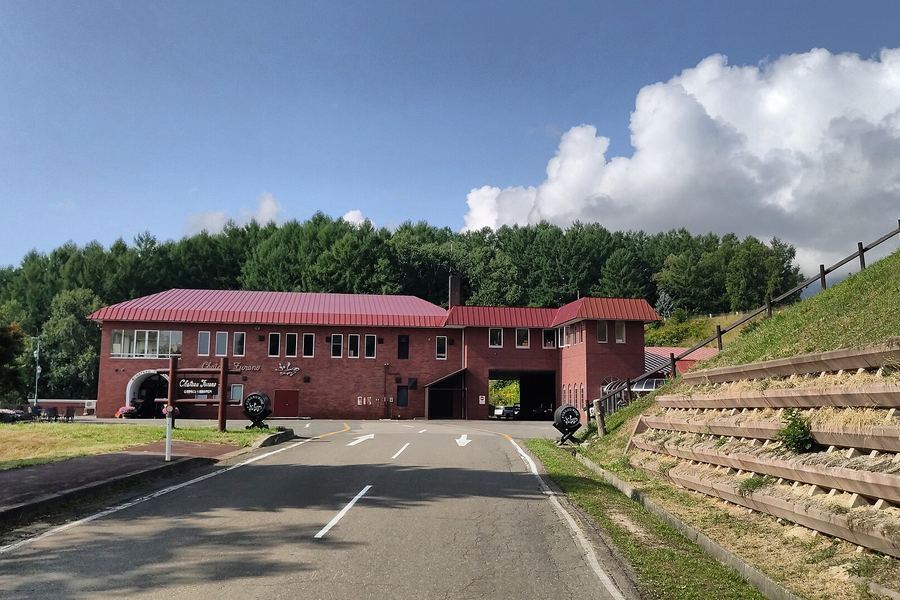 森を背にした、赤い屋根と赤れんがの2階建ての建物。入口に「Chateau Furano」の看板と樽のオブジェ。手前にゆるくカーブする道路と、右手に木の土留めのある緑の斜面