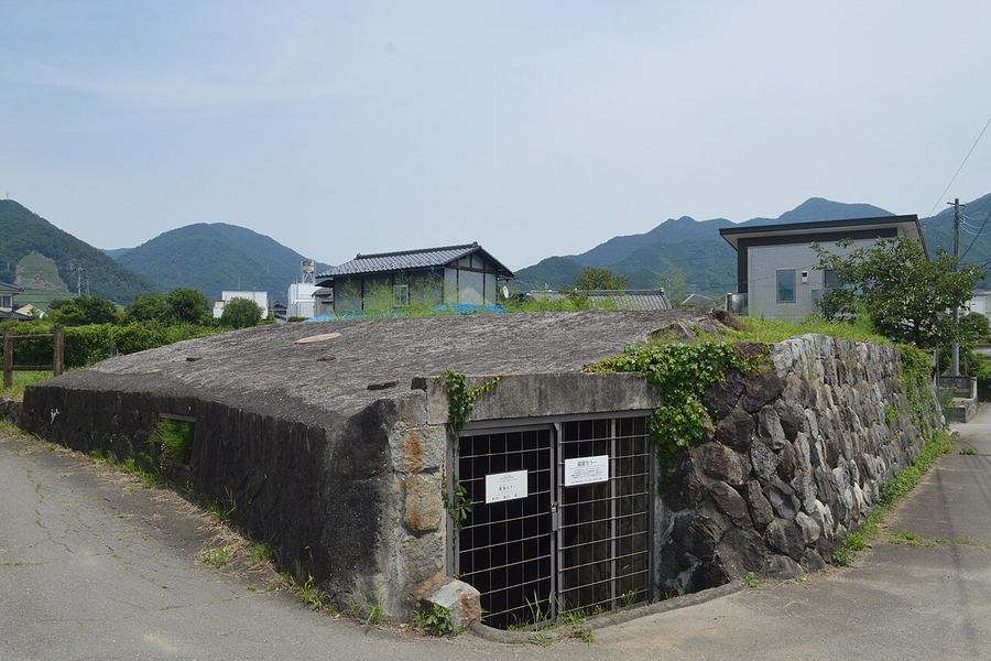 石積みの壁に挟まれた、れんがとコンクリートの半地下の貯蔵庫。正面に白い鉄格子の扉が二つ並び、屋根の上には草が生えている。背後に民家と緑の山並み