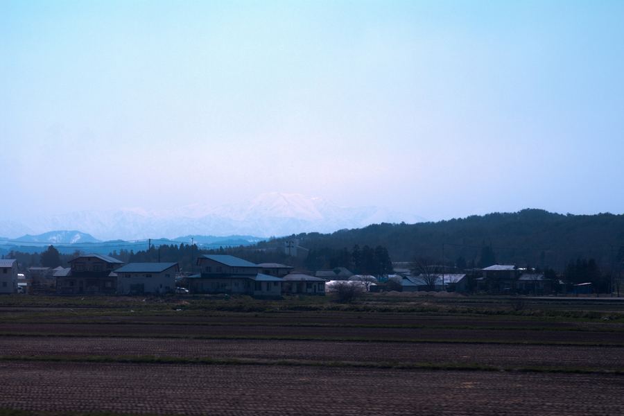 手前に耕された畑が広がり、その奥に家々と黒い森の丘が並ぶ。霞んだ空の向こうに、雪をかぶった山並みが白く浮かんでいる