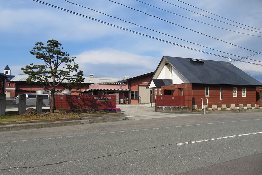 青空の下、道路沿いに赤茶色の外壁の建物が並ぶ。右手には黒い屋根に白い妻壁の大きな切妻の建物、左手には松の木と赤茶色の塀がある