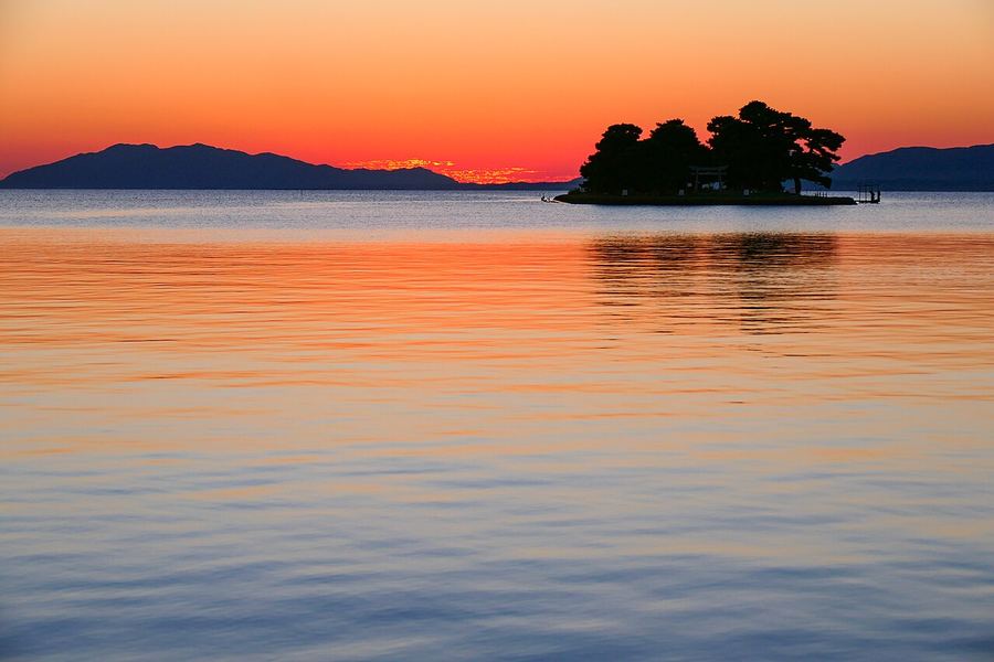 夕焼けで橙色に染まった静かな湖面。遠くの山並みの手前に、木の茂った小さな島が浮かんでいる