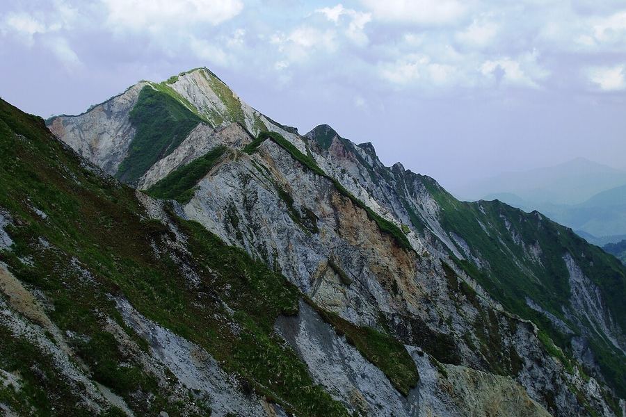 緑の草に覆われた険しい山の稜線。白っぽい岩肌の崖が谷へ落ち込み、奥に山並みがかすんでいる