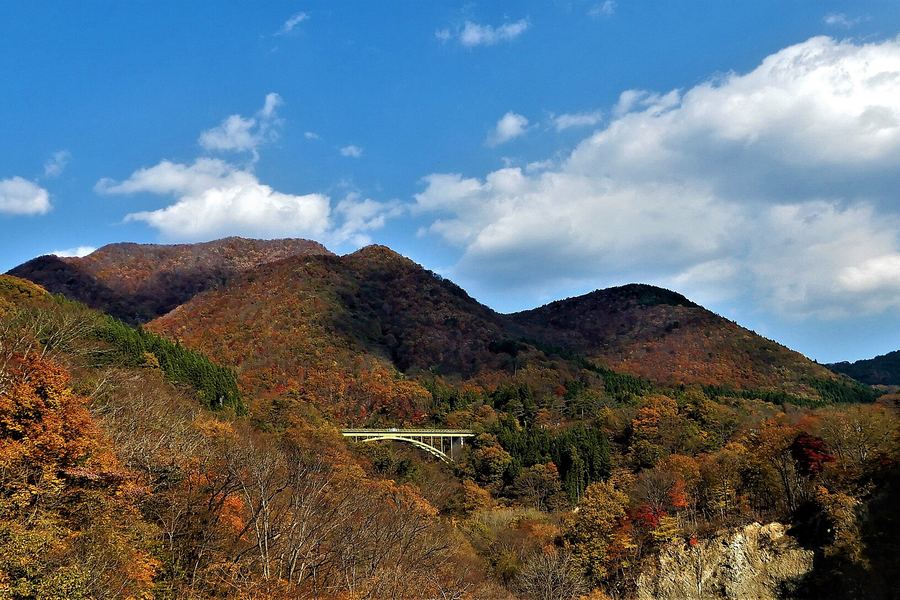 赤や茶色に色づいた木々に覆われた山並み。谷あいに白いアーチ橋が架かり、空には白い雲が浮かんでいる