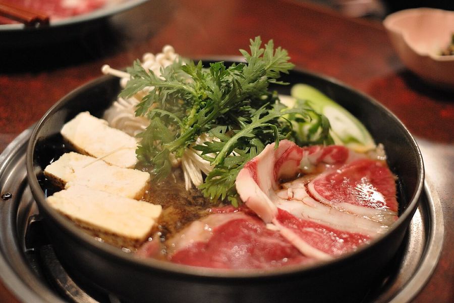 鉄鍋の中で、霜降りの牛肉、焼き豆腐、春菊、えのき、ねぎが割り下に浸かっているすき焼き