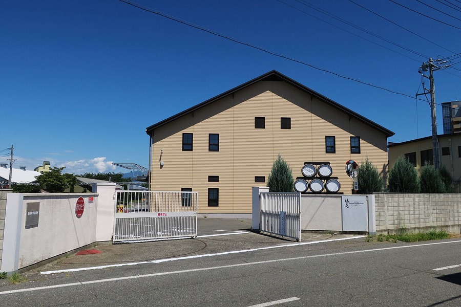 青空の下、門の奥に建つ切妻屋根の醸造棟と、積み上げられた樽の飾り